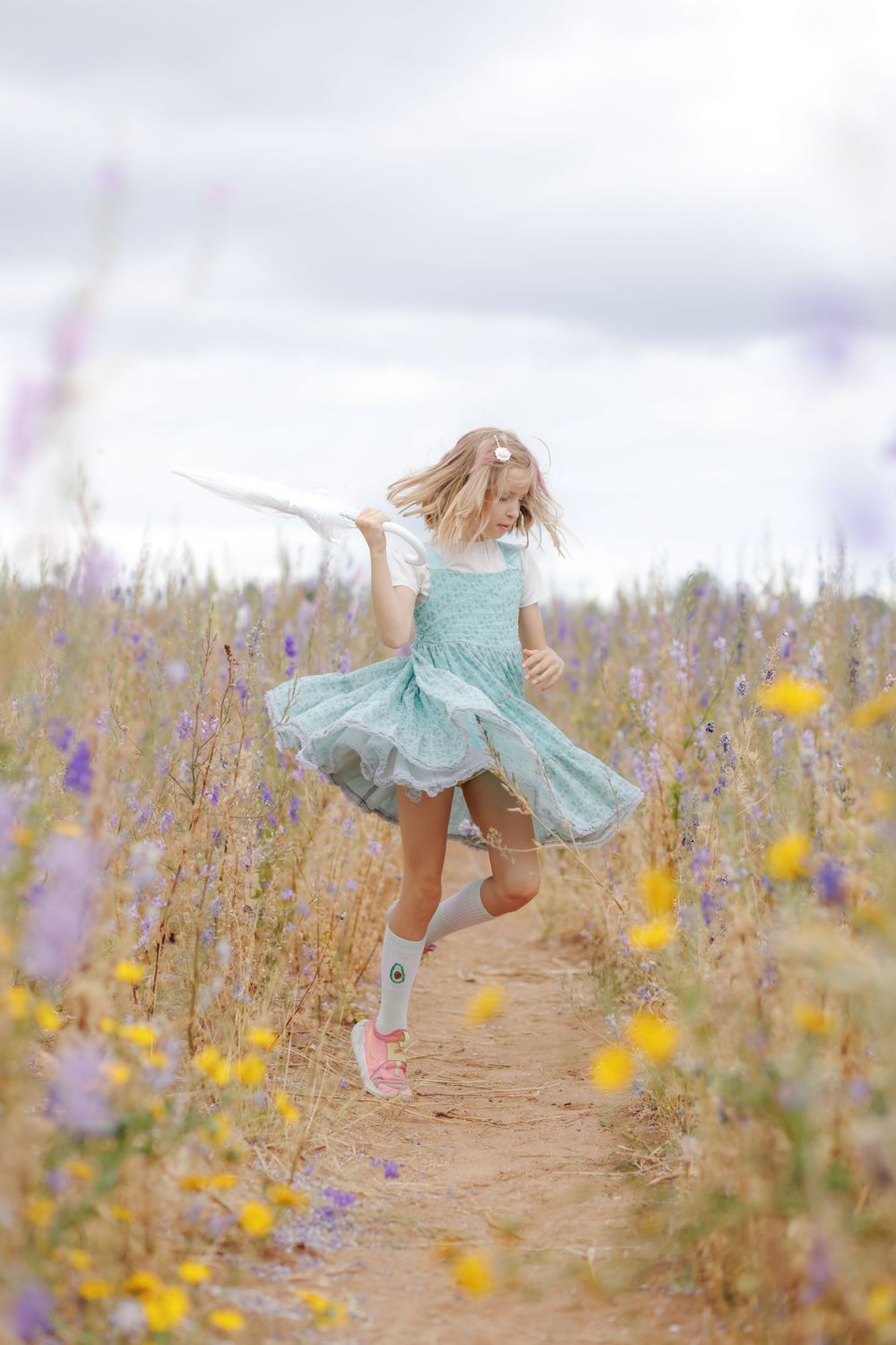 Young girl in teal dress twirling on a wildflower field path with purple and yellow blooms under cloudy sky.