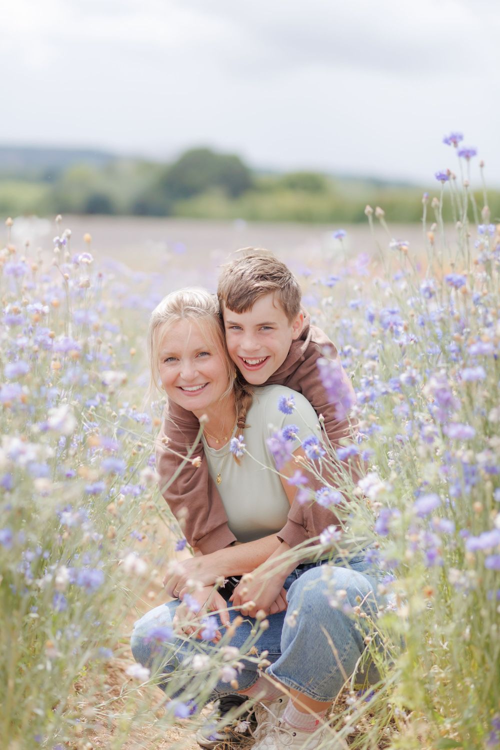 A woman and teenage boy embrace and smile in a purple wildflower field on a cloudy day.