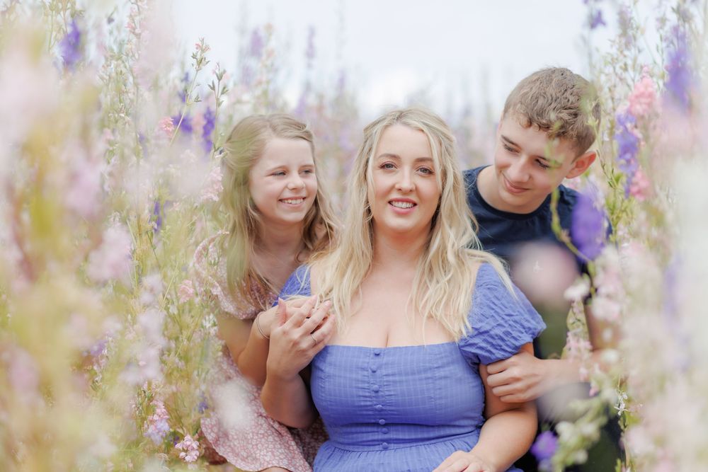 A woman in a blue dress poses with two children among blooming purple and pink wildflowers in a dreamy outdoor setting.