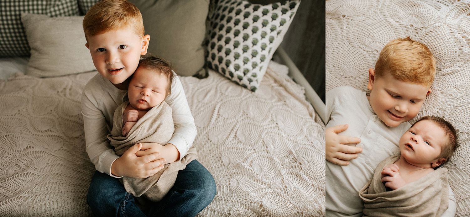 A smiling red-haired toddler boy gently holds his sleeping newborn sibling on a cozy cream knit blanket.