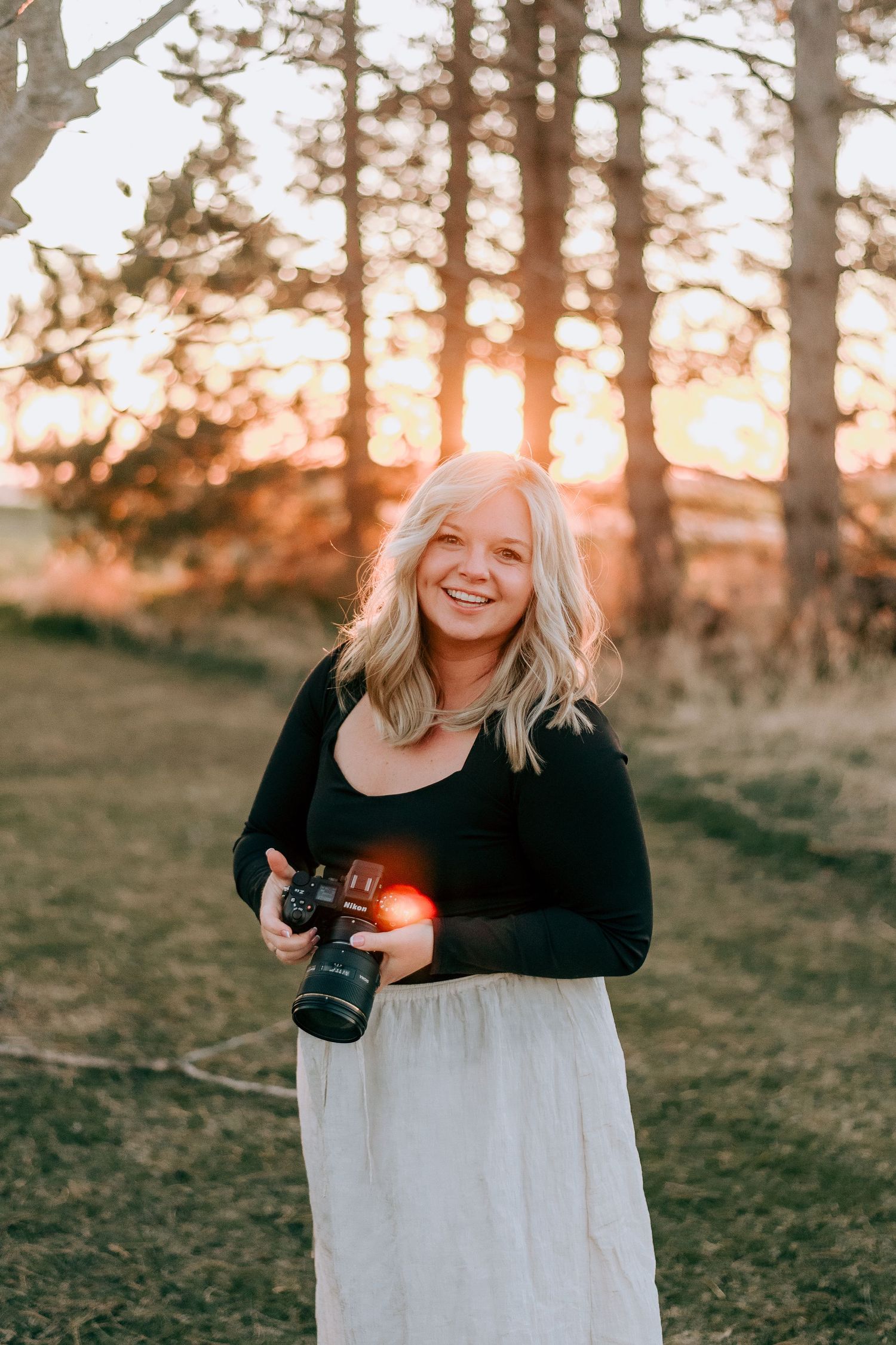 Smiling blonde woman holding a camera outdoors at golden hour with trees and sunset in background.