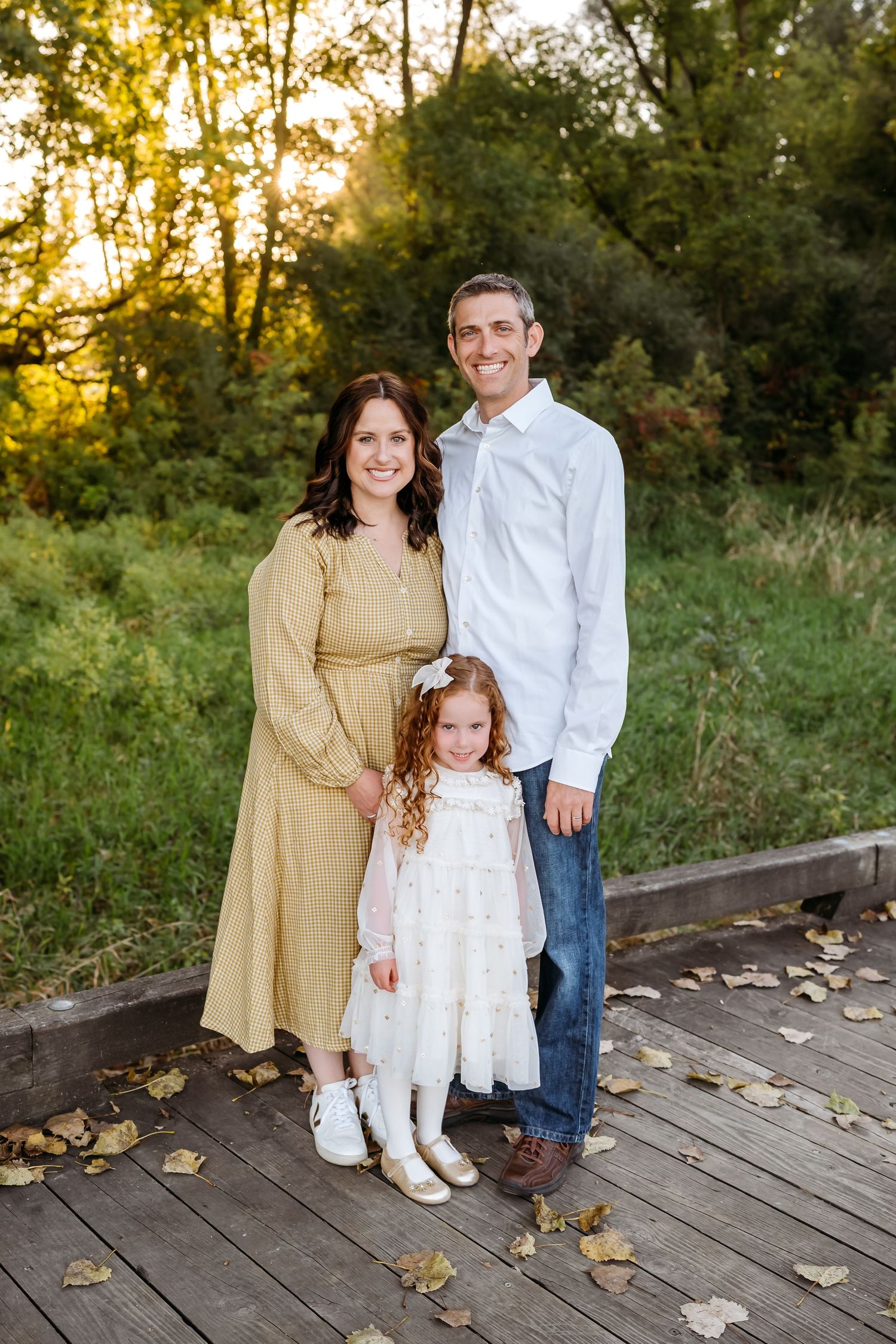 Family of three posing on wooden bridge surrounded by autumn foliage at golden hour sunset.