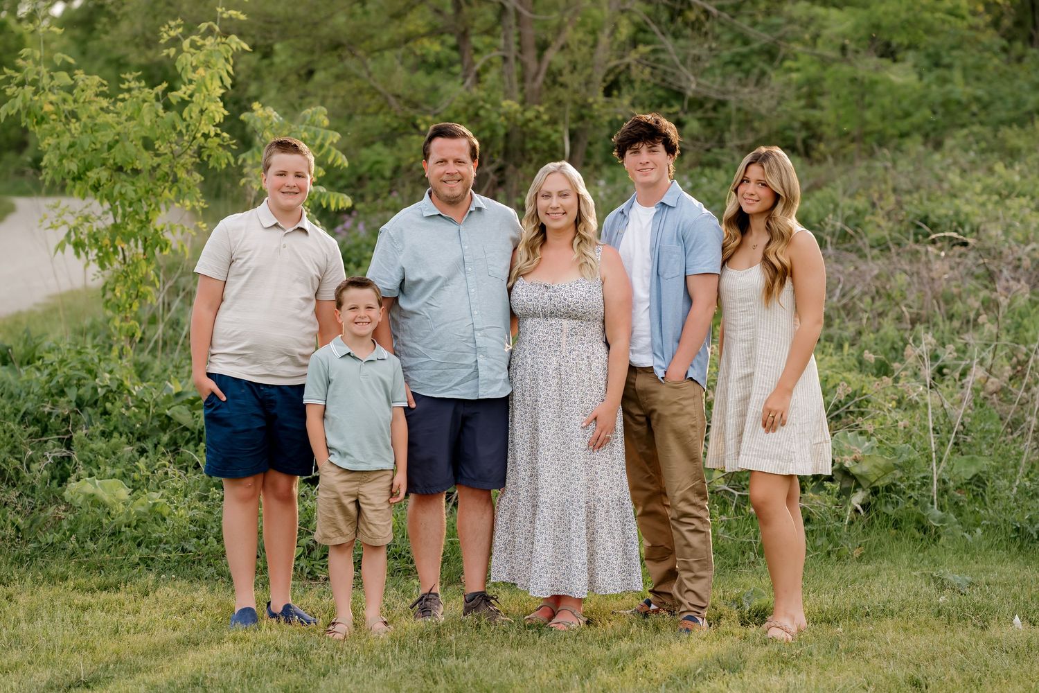 Family of six smiling outdoors in a green park setting, dressed in coordinated casual outfits for a portrait photo.