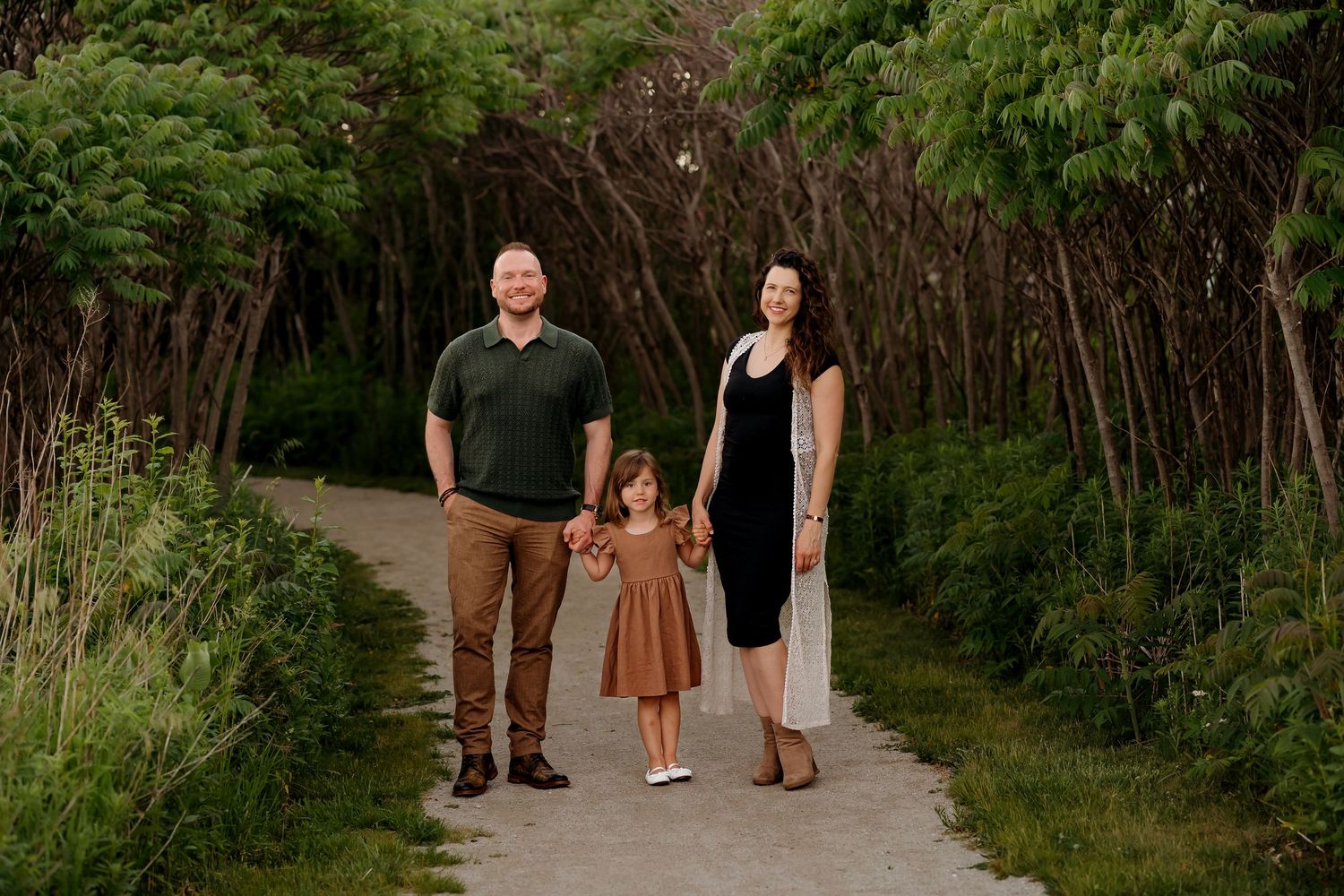 Family of three holding hands on a tree-lined path, parents in casual attire with young daughter in brown dress.