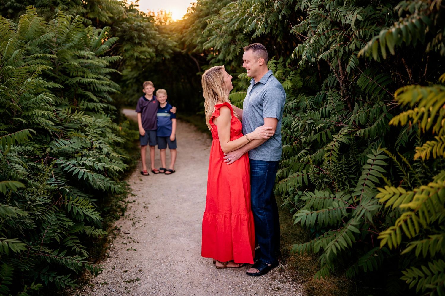 Couple in red dress and blue shirt share a romantic moment on a garden path at golden hour.