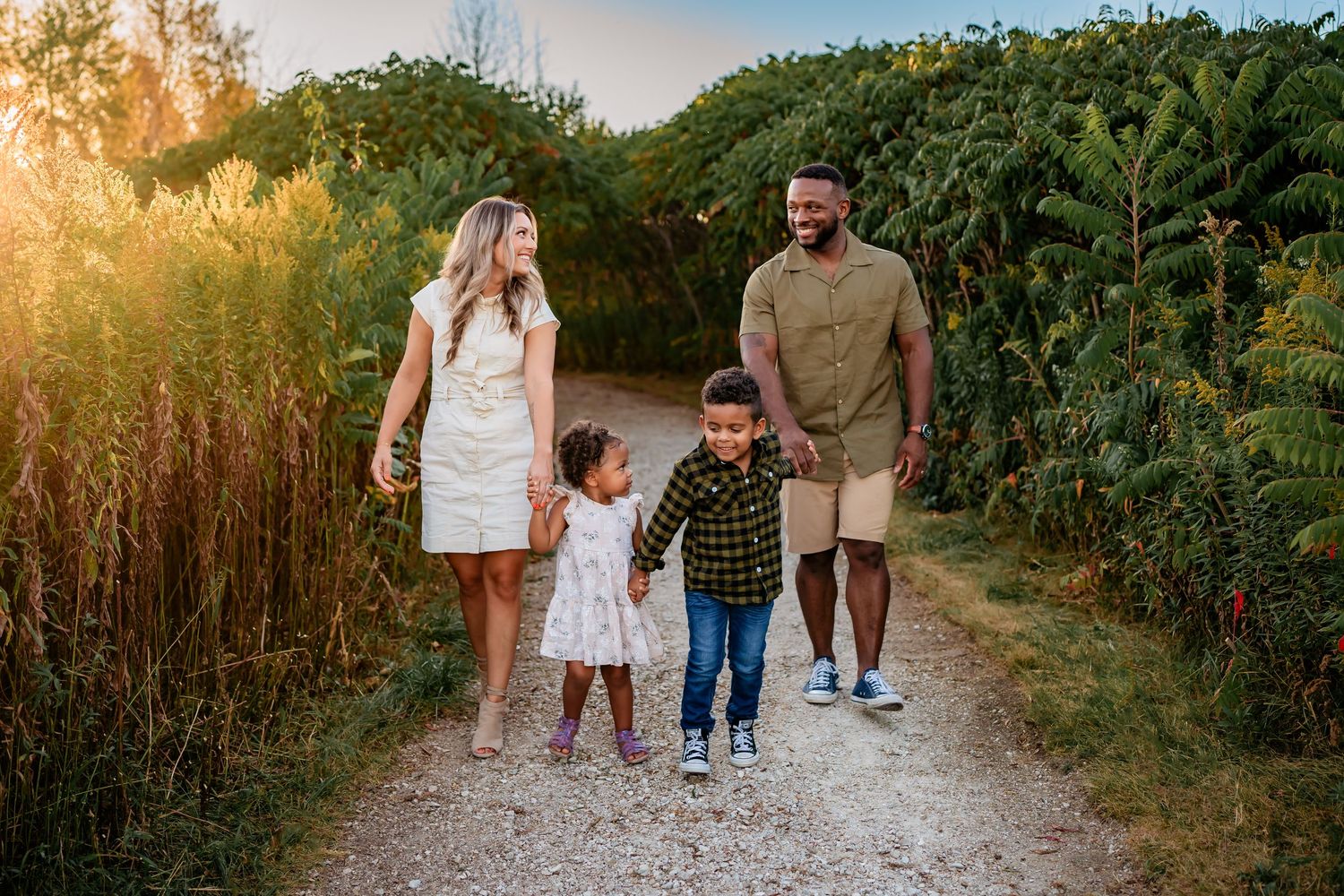 Family of four walking on a sunny garden path, parents holding hands with two young children among lush green trees.