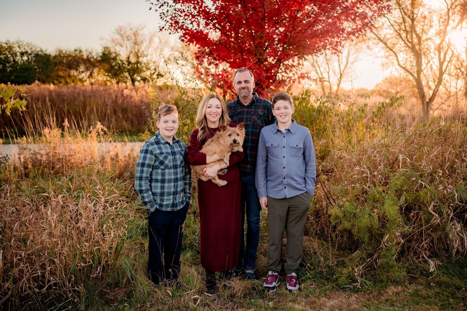 Family of four with a dog posing outdoors near a vibrant red tree during golden hour in autumn.