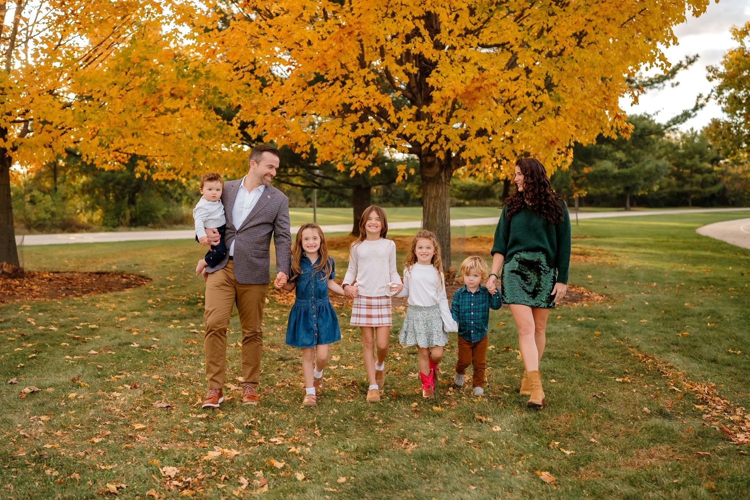 Family of six poses under vibrant yellow autumn tree on green lawn during fall season outdoor photo session.