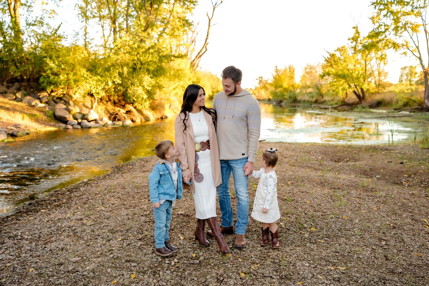 Family of four posing outdoors near a creek surrounded by golden autumn trees during a fall photo session.