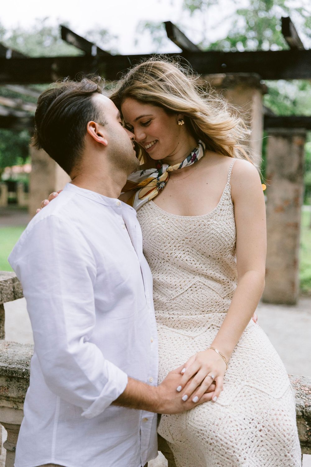 A couple shares a romantic moment outdoors, man in white linen shirt kissing woman in cream crochet dress.