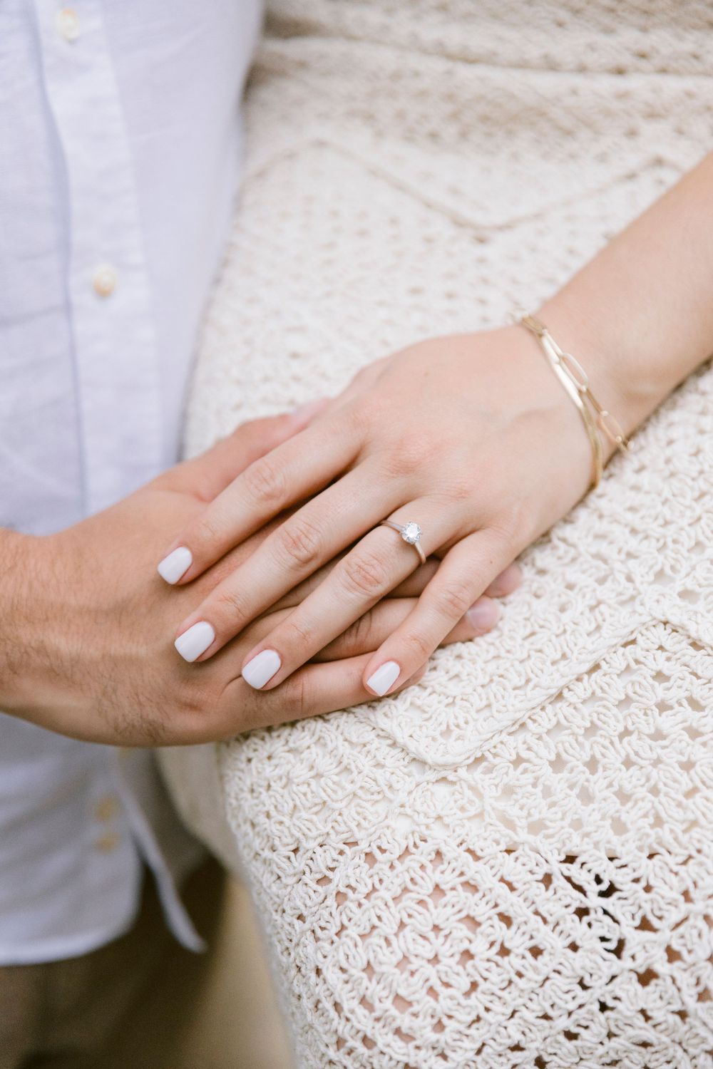 Close-up of engaged couple holding hands, woman wearing diamond ring, gold bracelet, white manicure, and crochet dress.