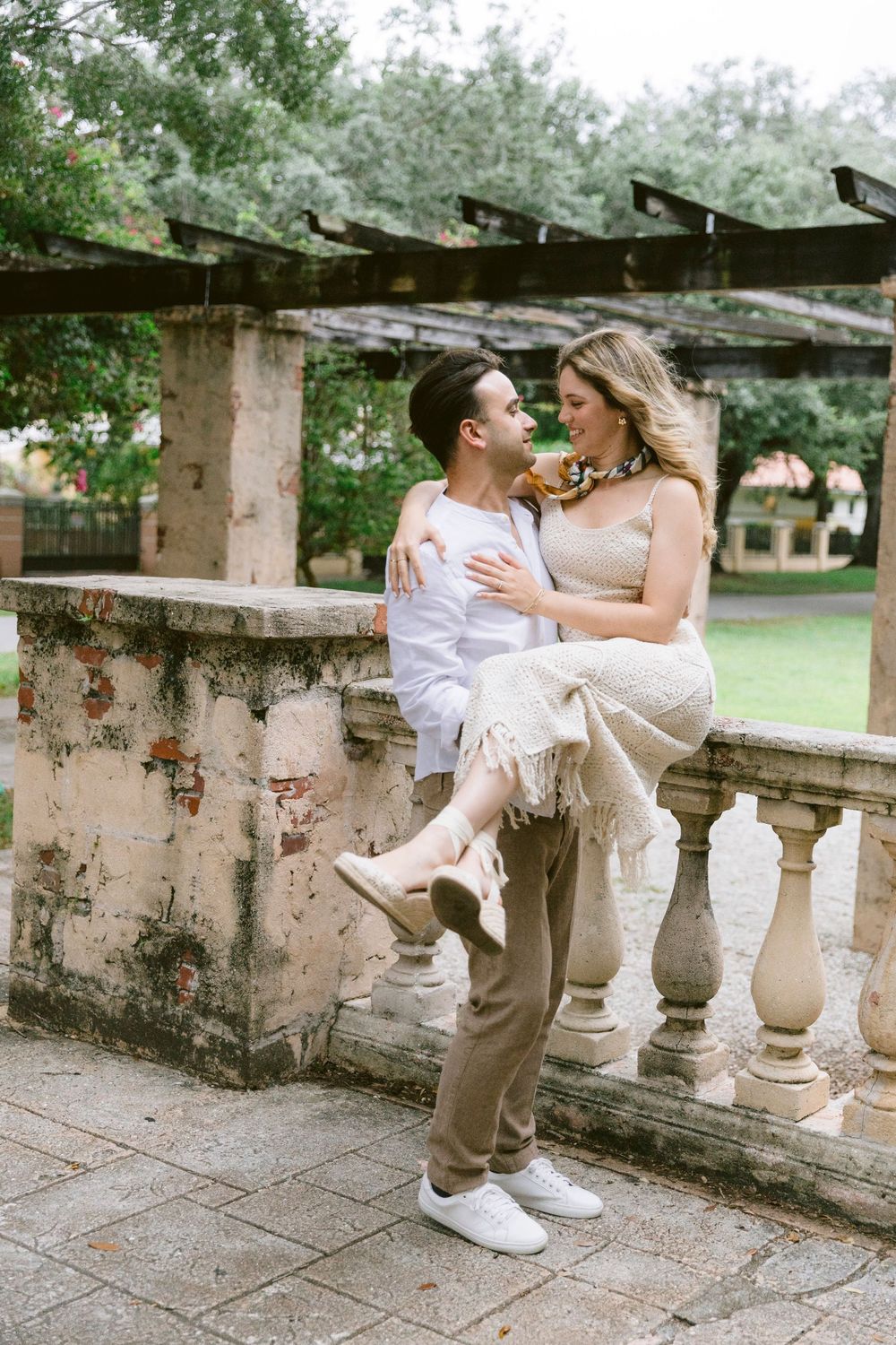 Romantic couple embracing on a stone balustrade in a lush garden, man holding woman as they gaze lovingly at each other.