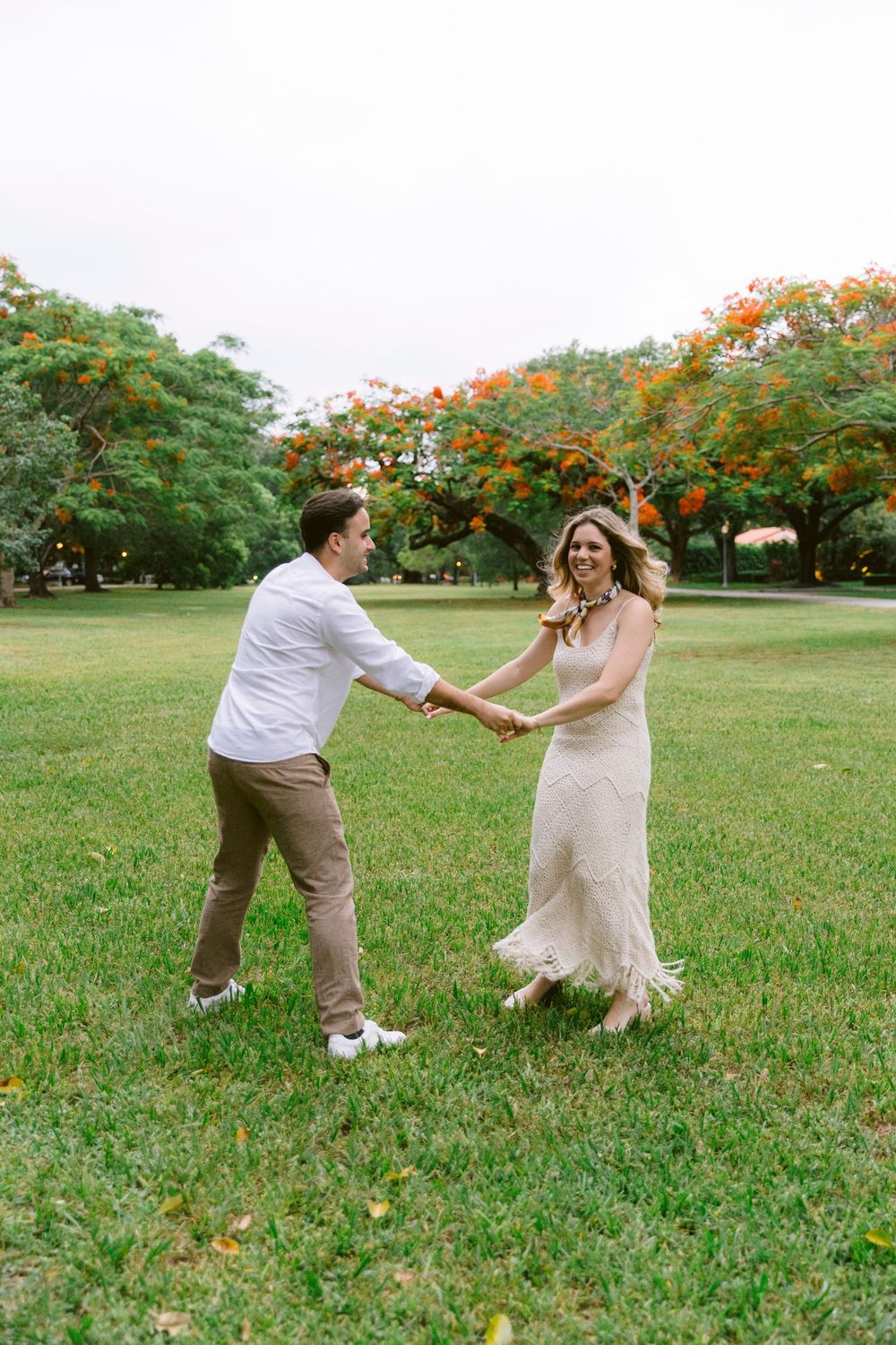 Couple dancing and holding hands on green grass lawn with vibrant orange flowering trees in background.