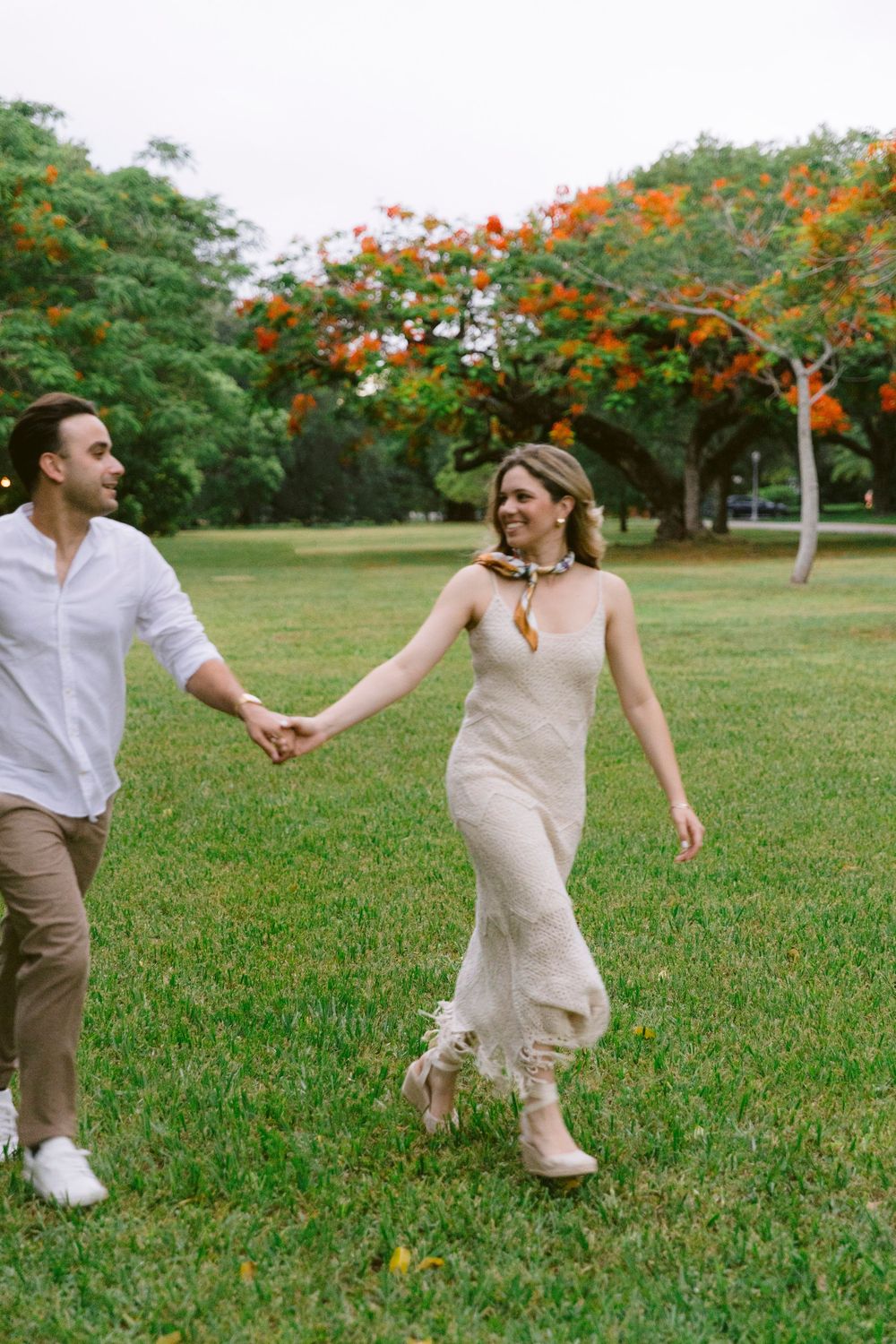 Couple holding hands walking in lush green park with orange flowering tree backdrop.