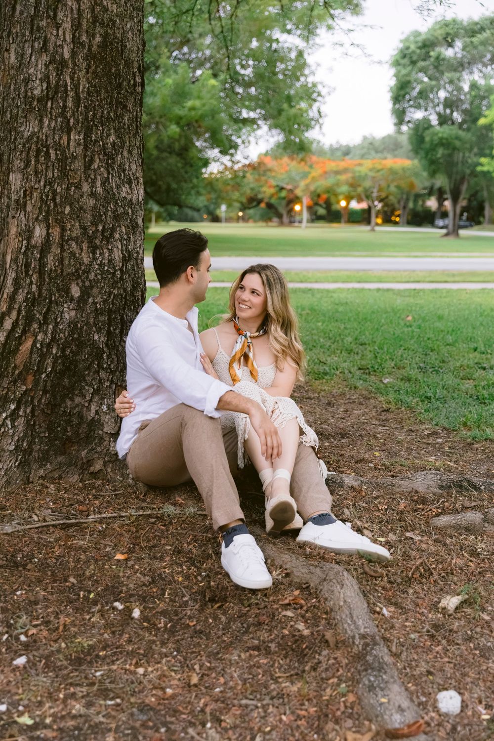 A couple sits together under a large tree in a lush green park, smiling at each other in casual neutral outfits.