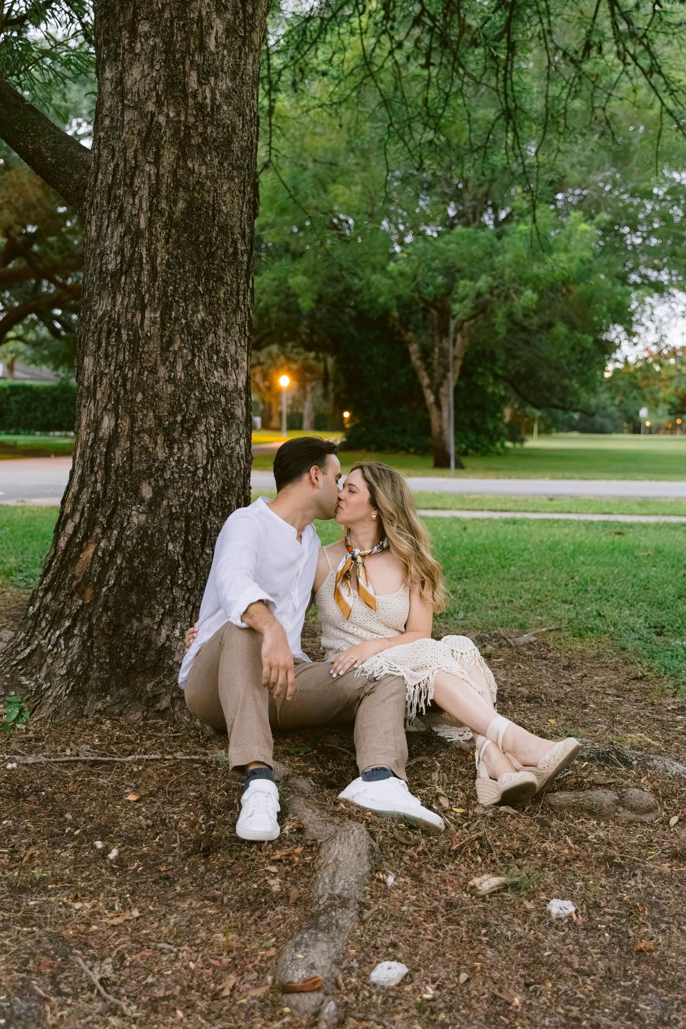 Couple sitting together under a large tree in a park, leaning in for a kiss at dusk.