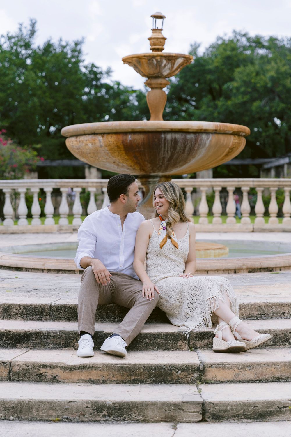 Couple sitting on fountain steps in a garden, sharing a romantic moment, dressed in casual elegant attire.
