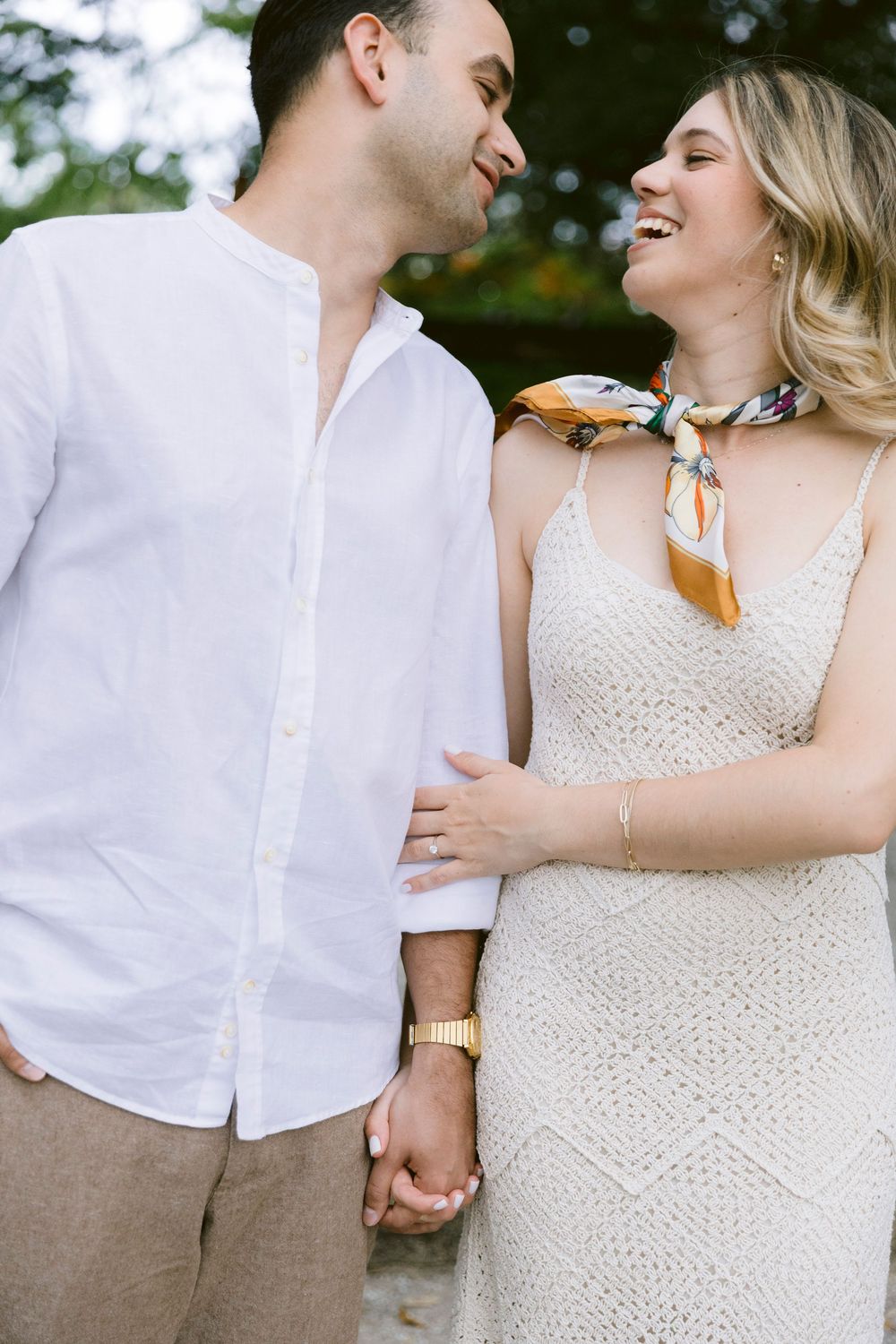 A smiling couple holds hands outdoors, woman in white crochet dress with scarf, man in white linen shirt.