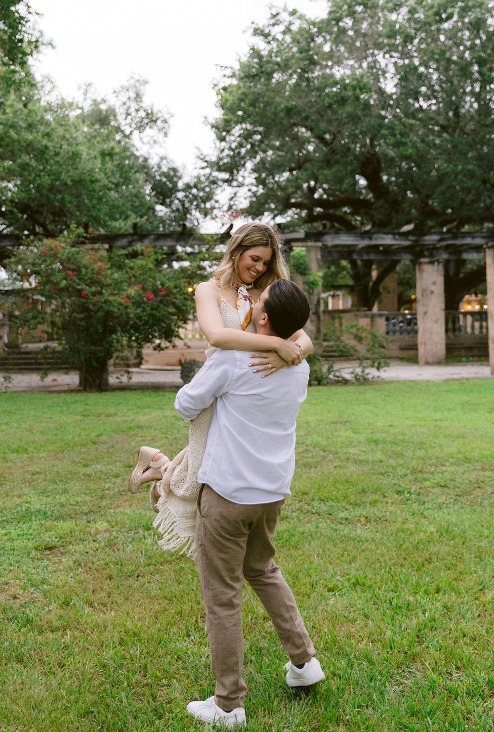 A man lifts and embraces a woman in a lush green park, both smiling joyfully during an outdoor engagement session.