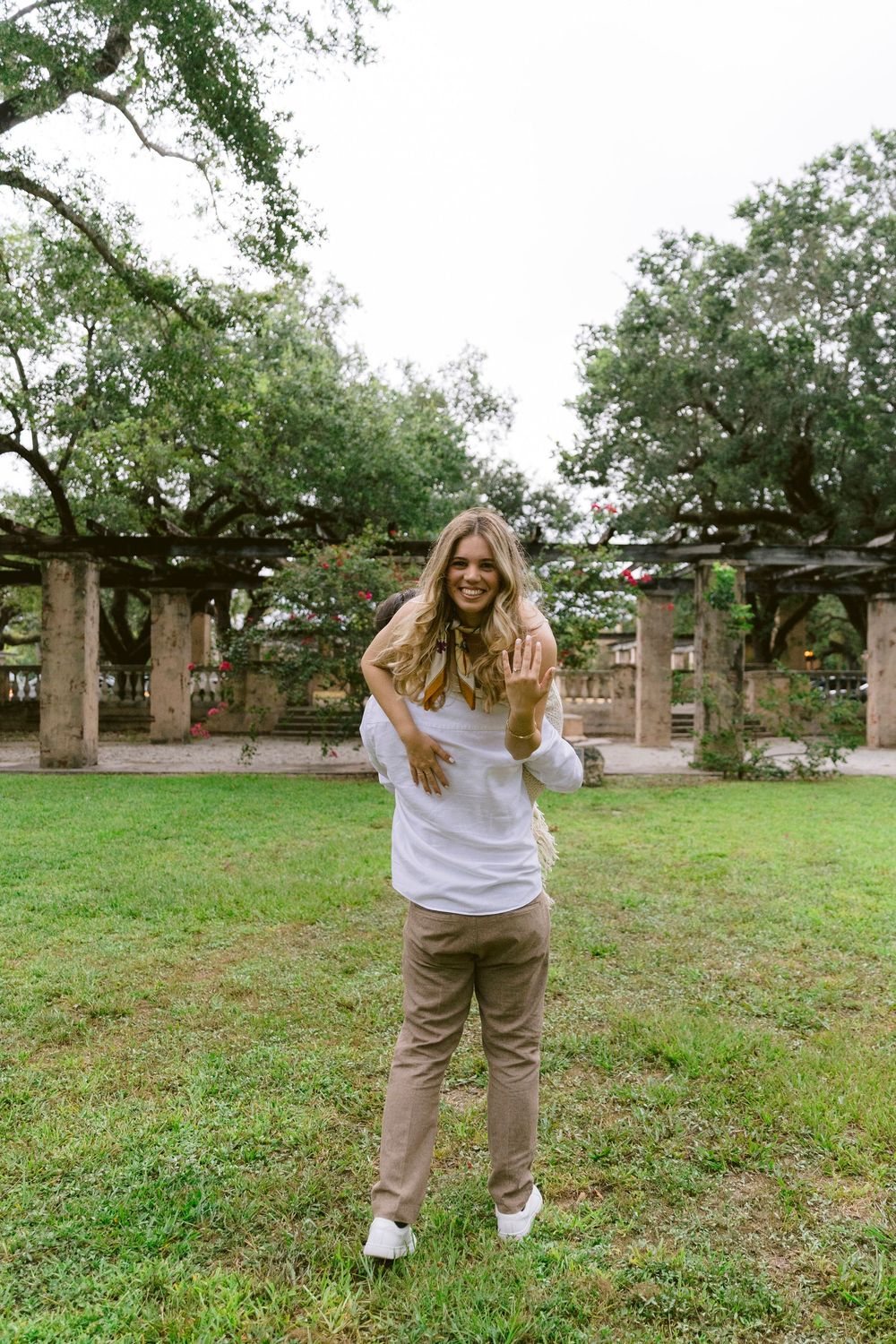A smiling woman in white top and tan pants poses playfully in a lush green park with stone pergola.