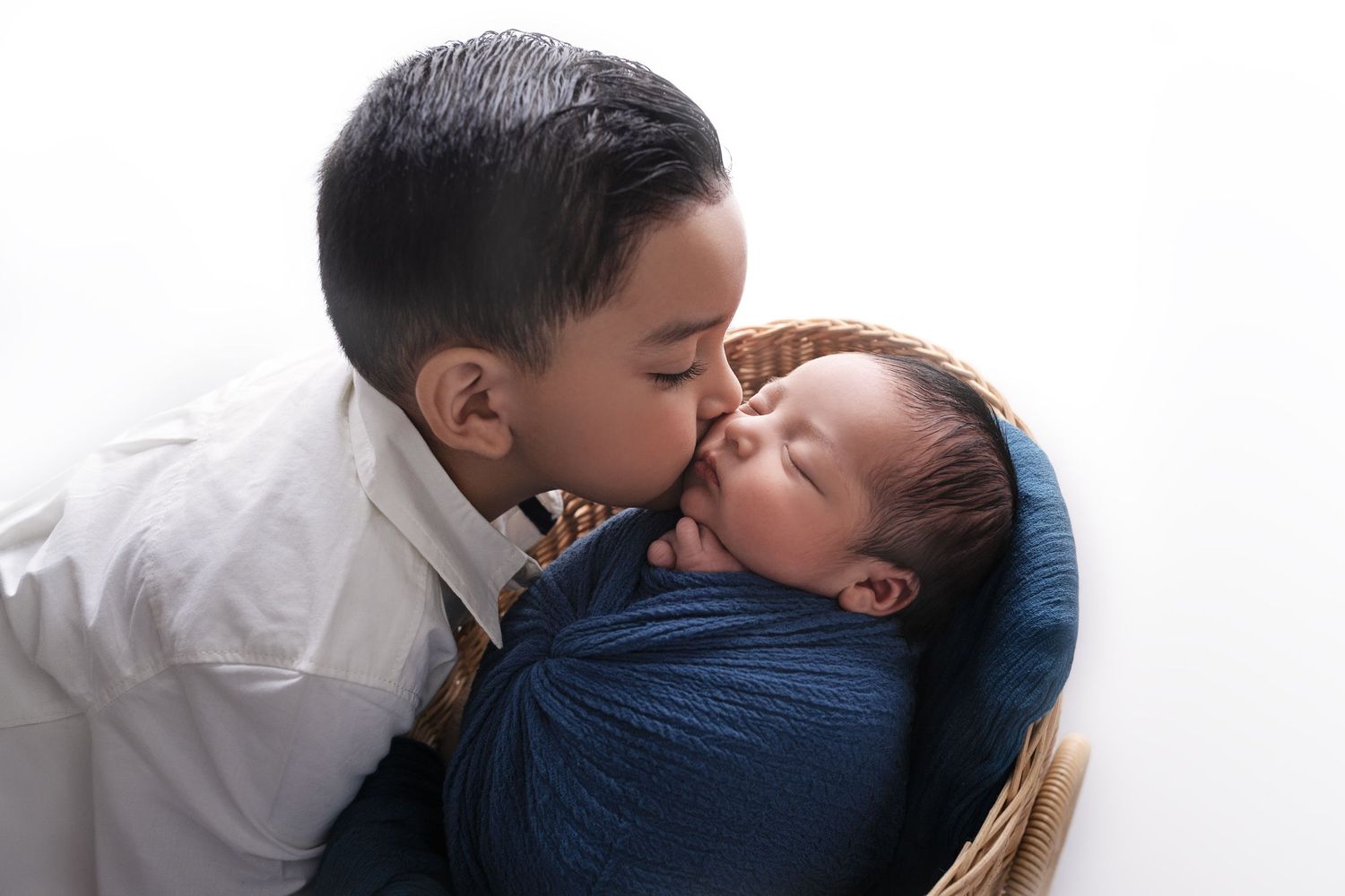 Older boy gently kisses newborn baby wrapped in blue swaddle in a basket on white background.