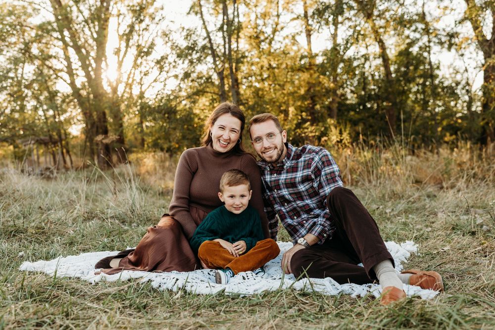 Family of three smiling on a blanket in a sunlit autumn field, parents embracing young boy in green sweater.