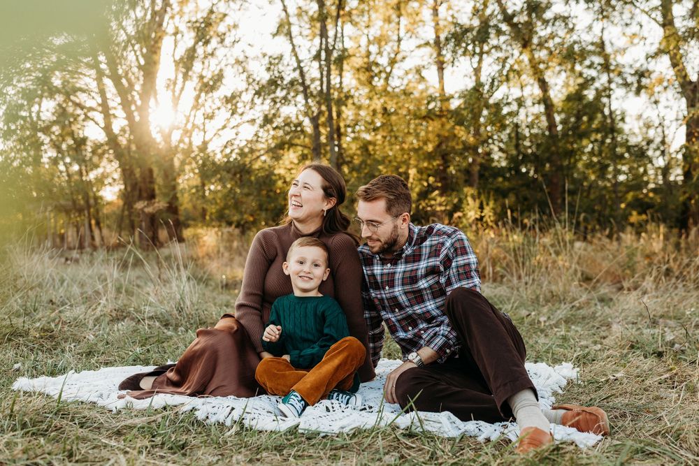 Family of three sitting on blanket in autumn field, parents smiling with toddler in green outfit, golden sunset.