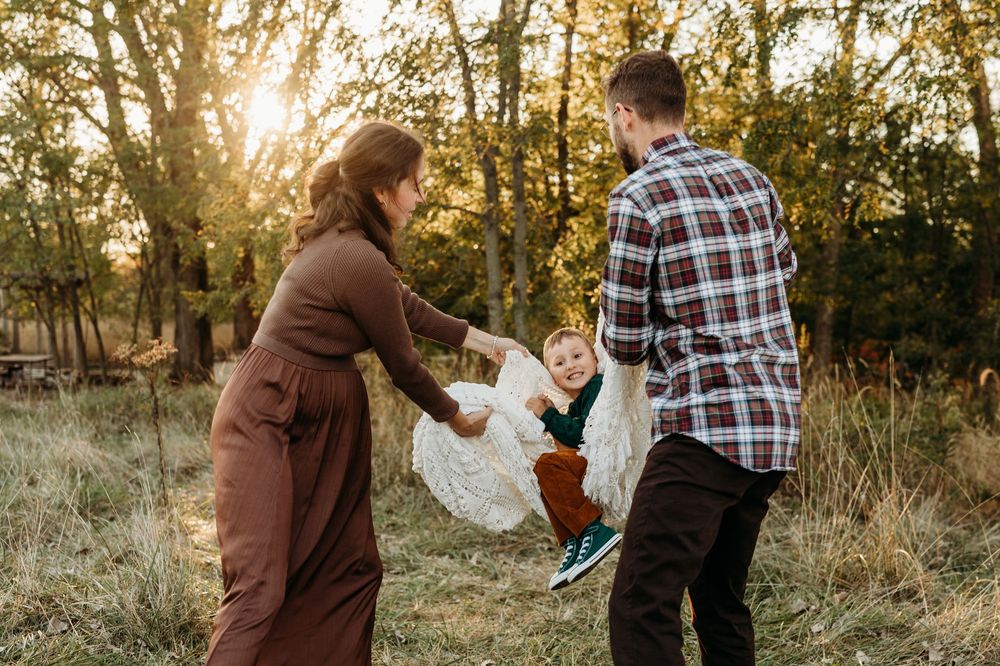 A young family swings their toddler between them in a sunlit autumn field surrounded by golden trees.