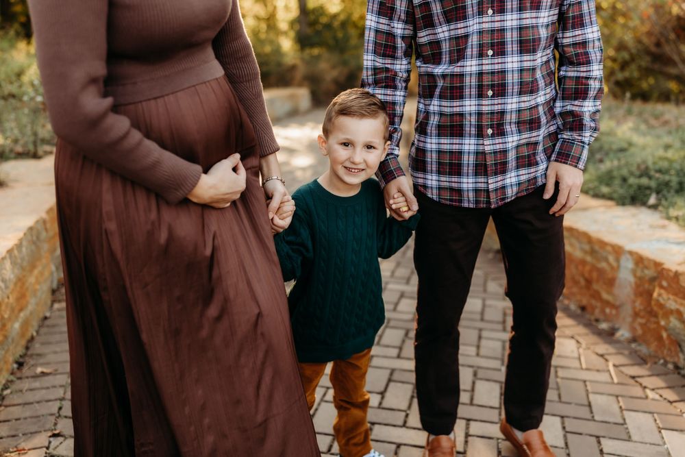 A young boy holds hands with his pregnant mother and father during a fall family portrait session outdoors.