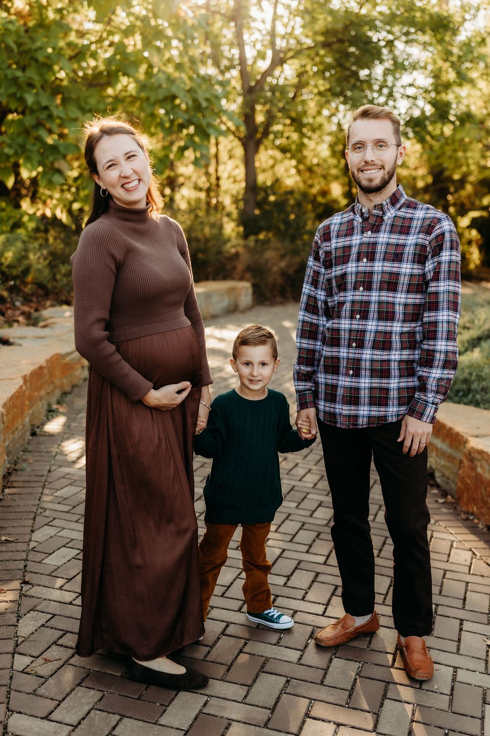 Pregnant woman in brown dress poses with husband in plaid shirt and young son on brick path in sunny park.