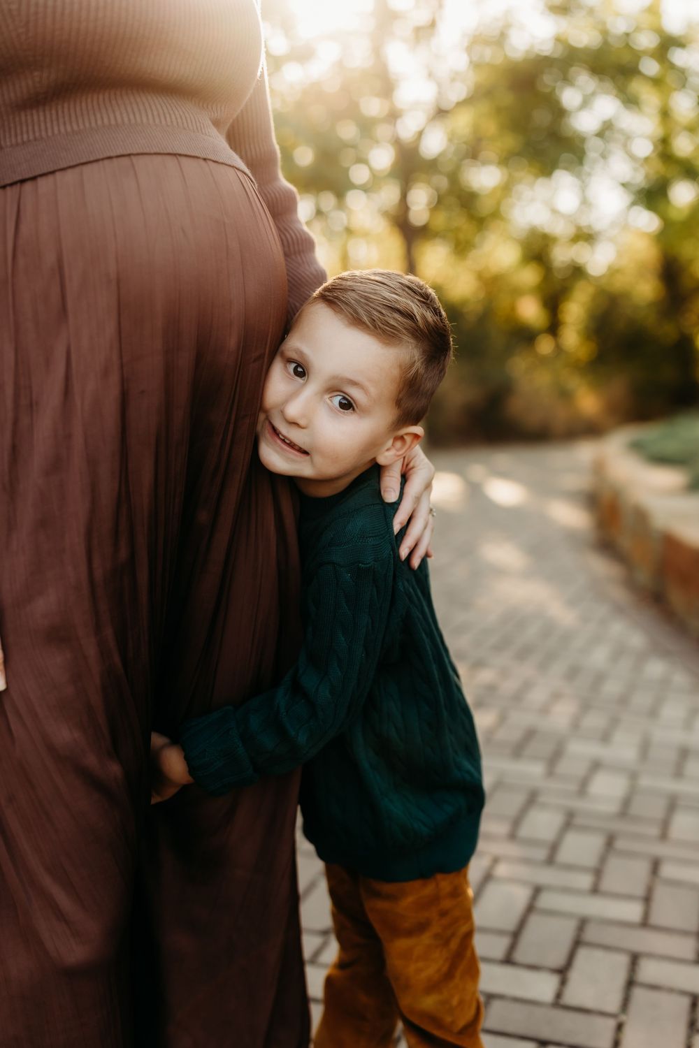 A young boy in a teal sweater hugs a pregnant woman's belly outdoors on a brick path in warm golden light.