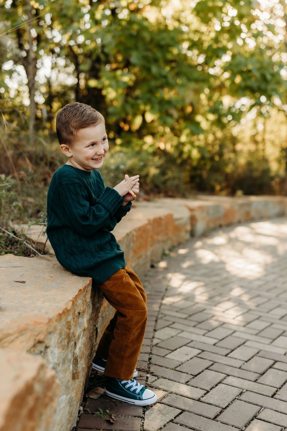 A smiling young boy in a green sweater and brown pants sits on a stone wall in an autumn park setting.