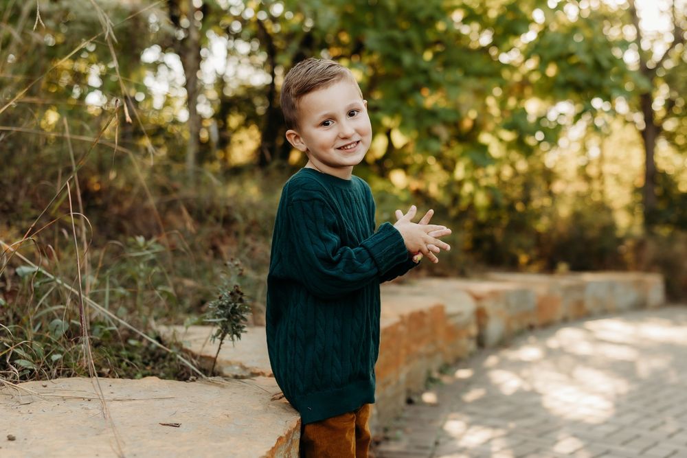Young boy in green sweater smiling and clapping hands outdoors in a park during autumn.
