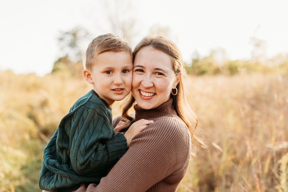 A smiling mother holds her young son outdoors in a sunlit field during a warm family portrait session.