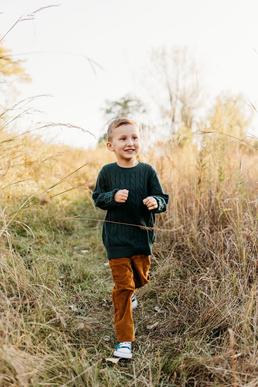 A happy young boy in a green sweater and brown pants runs through a golden autumn field outdoors.