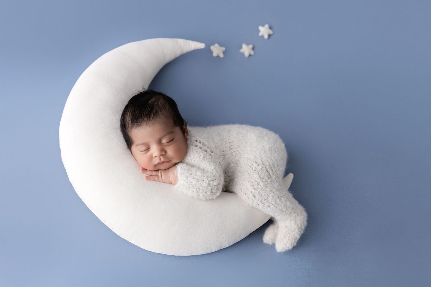 Newborn baby sleeping peacefully on a white crescent moon prop with stars on a soft blue background.