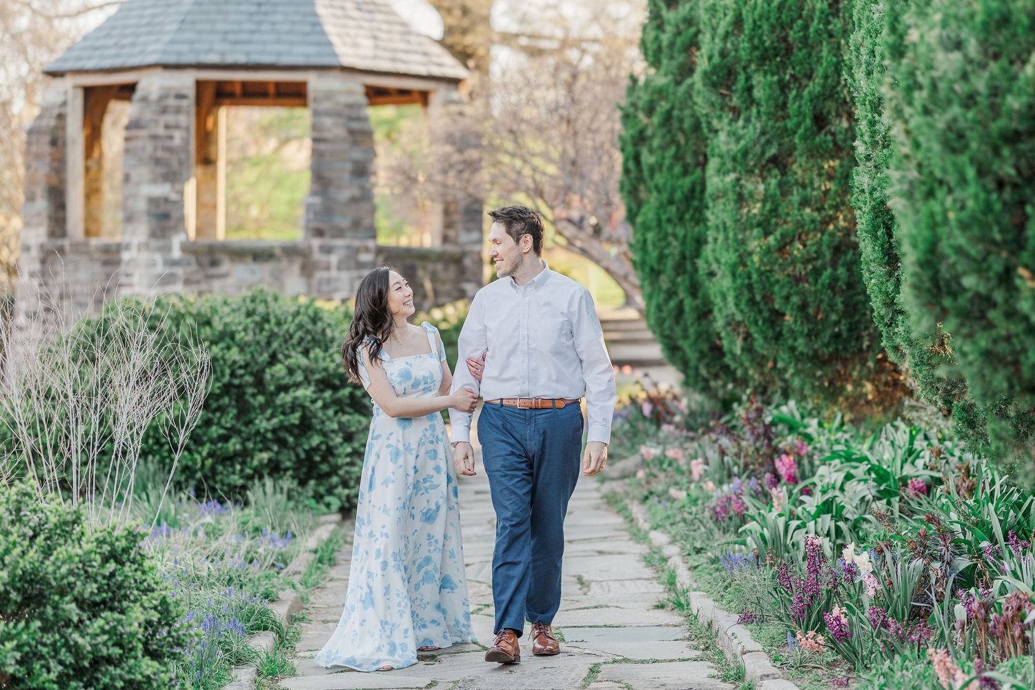 Couple walks hand-in-hand through a blooming garden path with stone gazebo and evergreen trees.