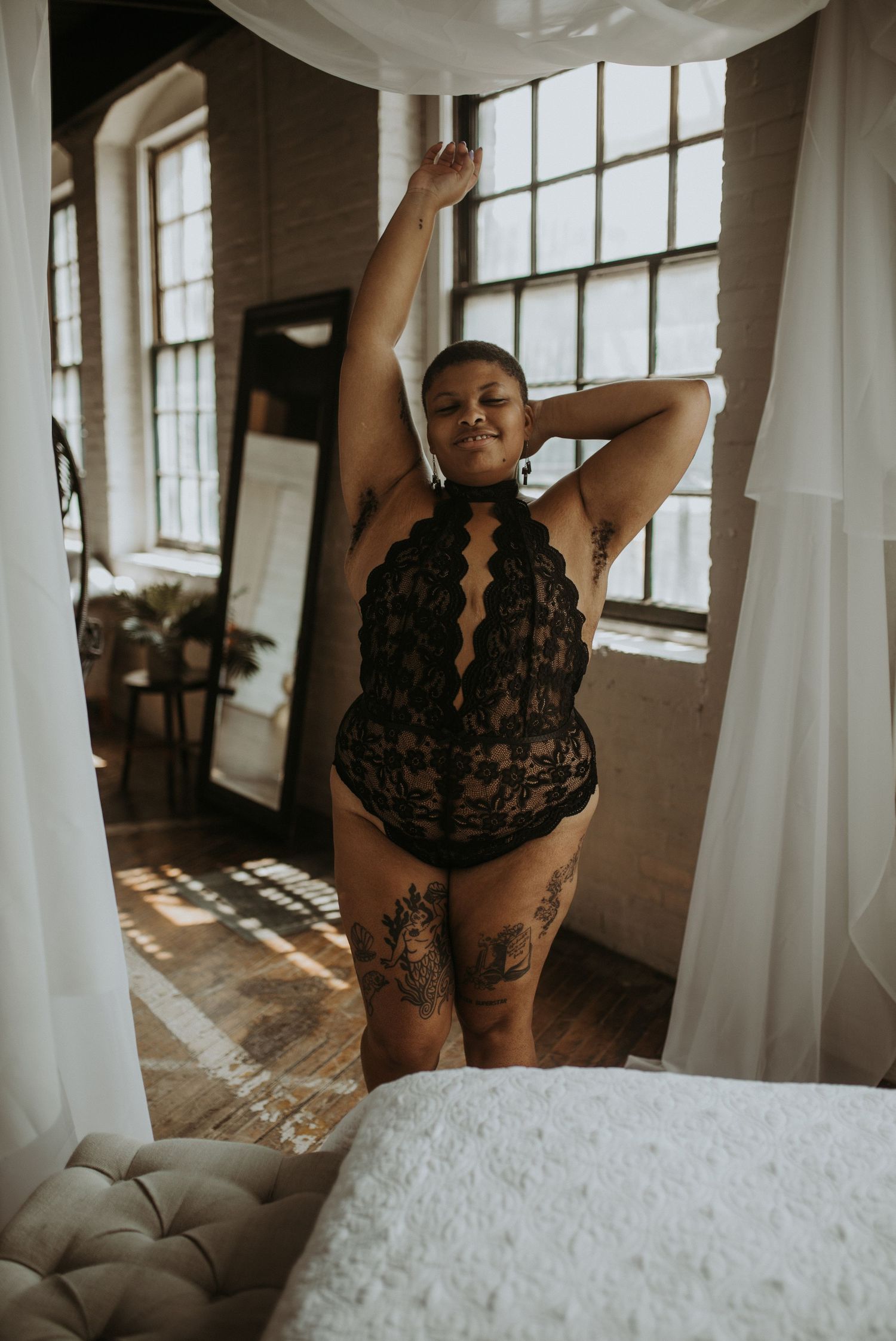 Laughing woman in purple lace top with tattoos sitting on white bed, wearing glasses, joyful expression.