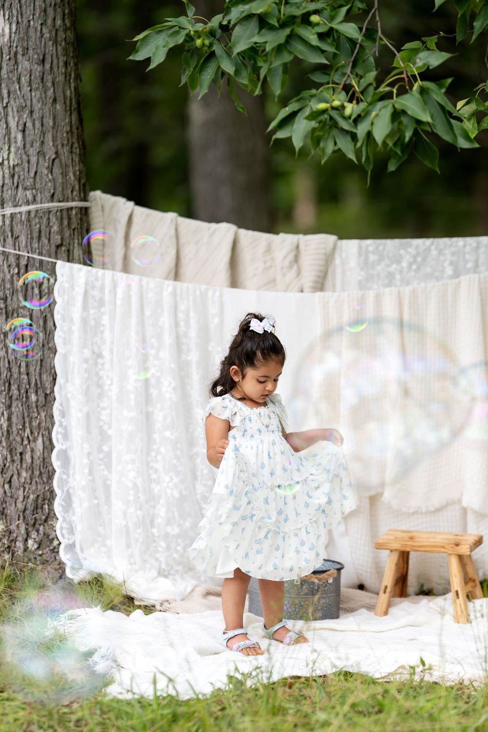 Young girl in white floral dress playing with bubbles outdoors near hanging linens and a tree.