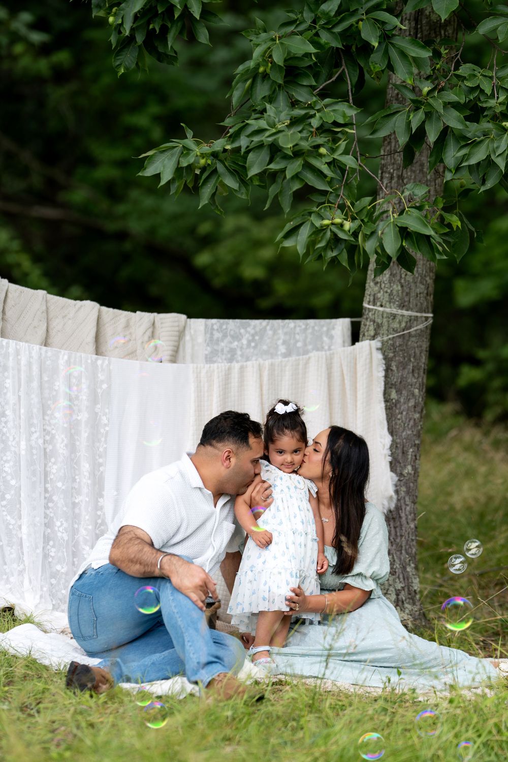 Family of three sitting on grass under a tree with white linens, parents kissing toddler girl surrounded by bubbles.