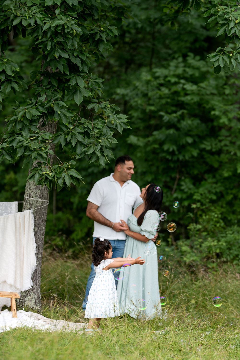 Family enjoying outdoor picnic with parents embracing while young daughter blows bubbles in lush green park setting.