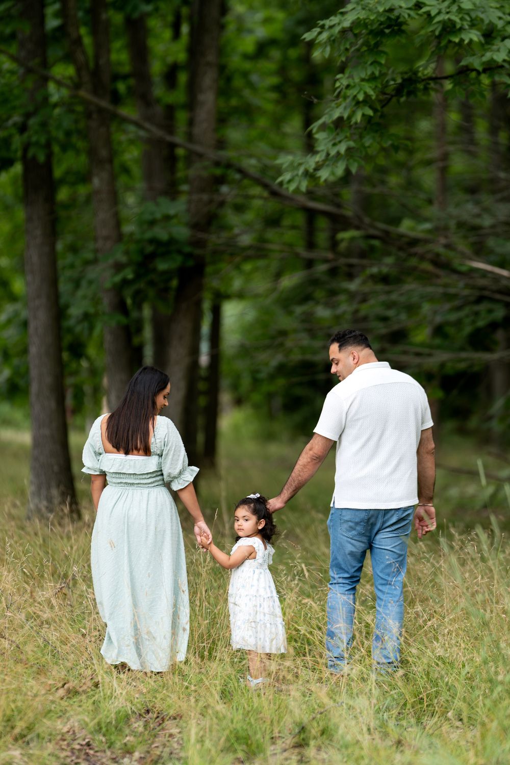 Family of three walking hand-in-hand through a lush green forest, parents in light blue dress and white shirt.