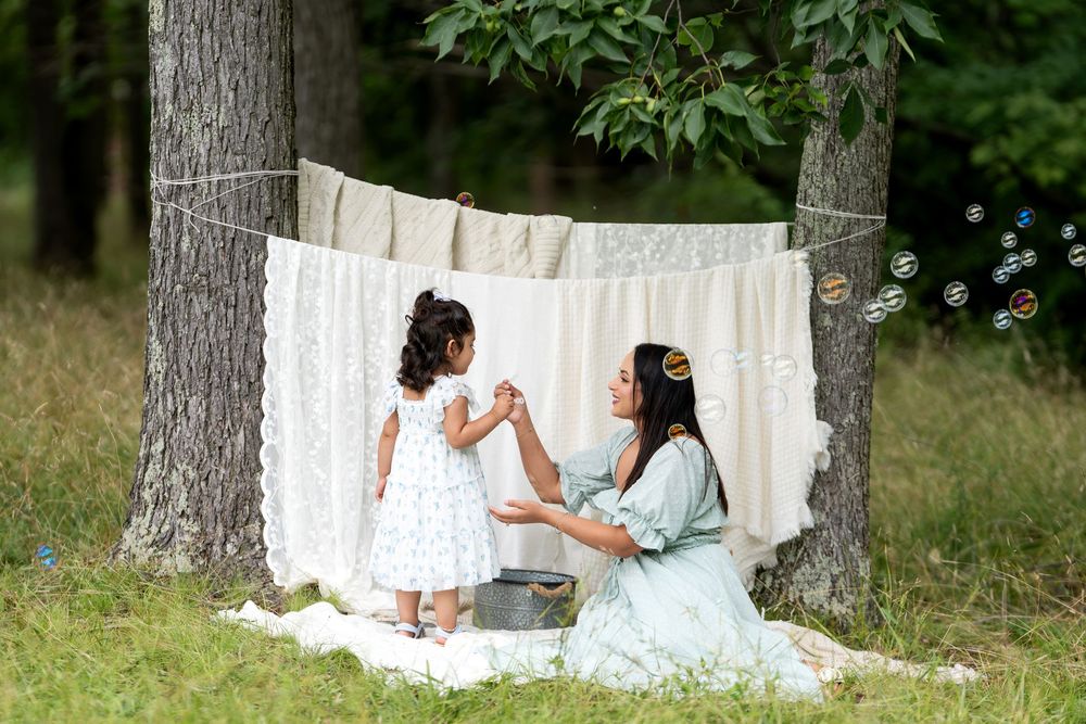 Mother and daughter playing with bubbles outdoors near lace fabric hung between trees on a sunny day.