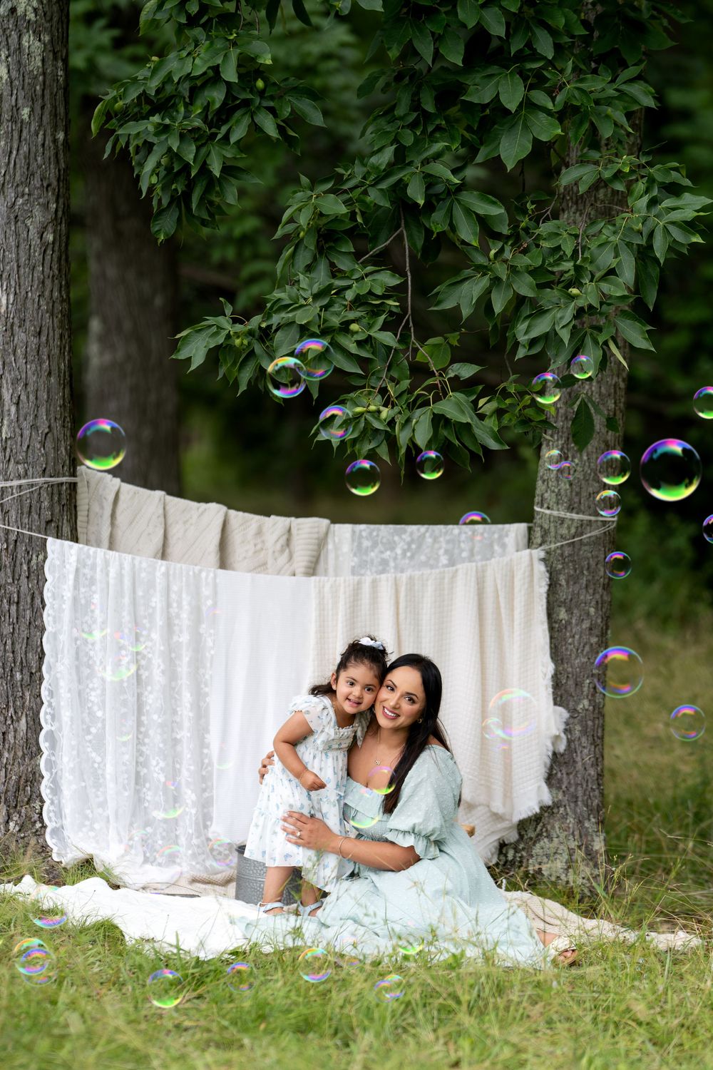 Mother and daughter sitting outdoors among soap bubbles, with white linens hanging between trees in a lush green park.