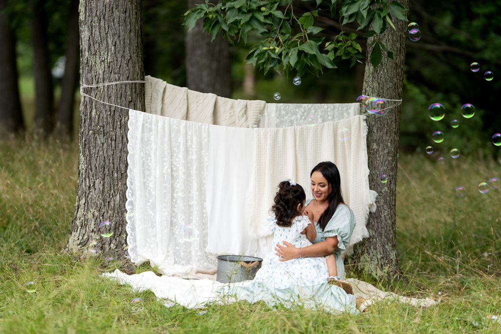 Two women sitting together outdoors under hanging white linens between trees, surrounded by floating bubbles in a serene setting.