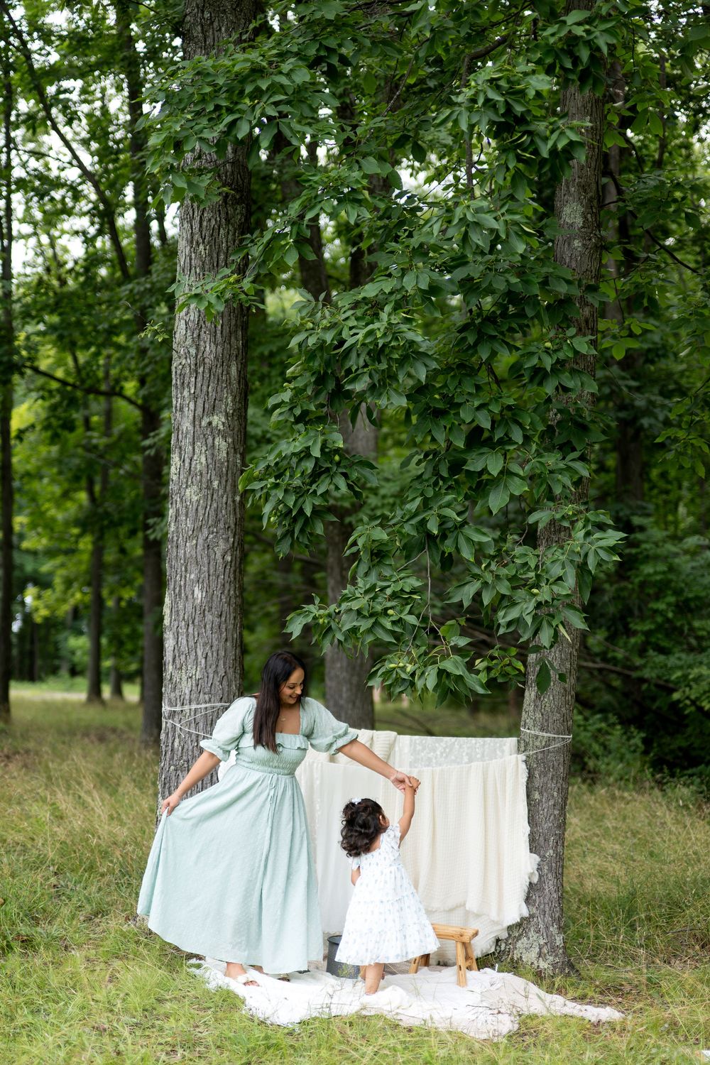 Mother and daughter in matching dresses dancing outdoors near linen draped between trees in a lush green forest.
