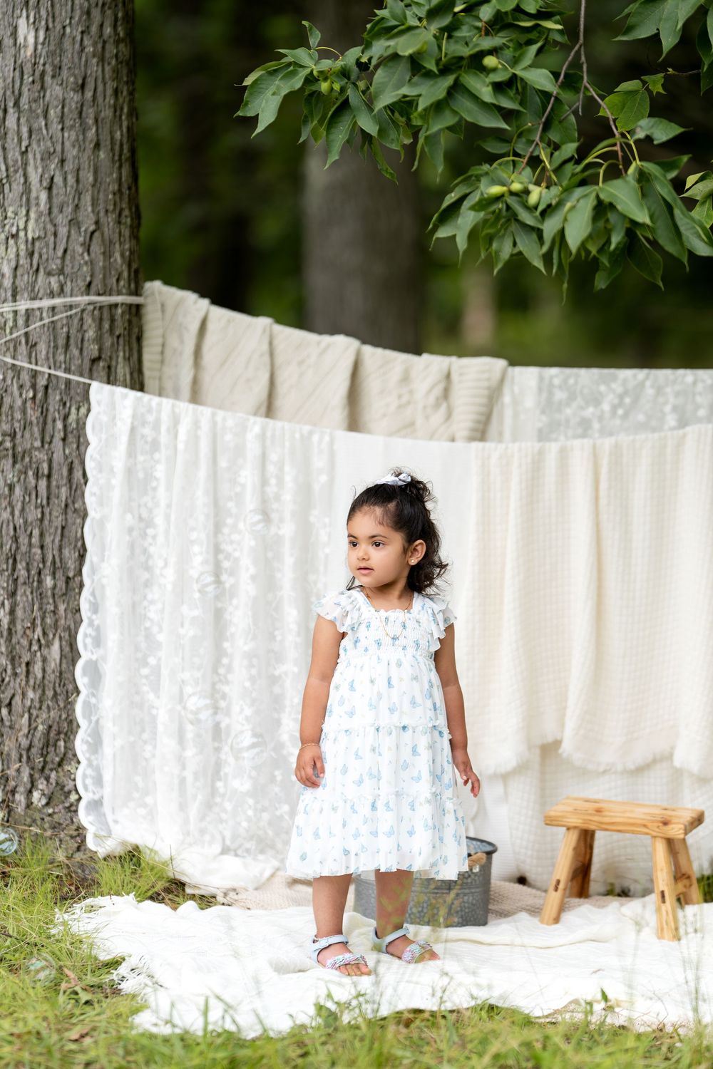Young girl in floral dress standing outdoors near hanging white linens on a clothesline between trees.