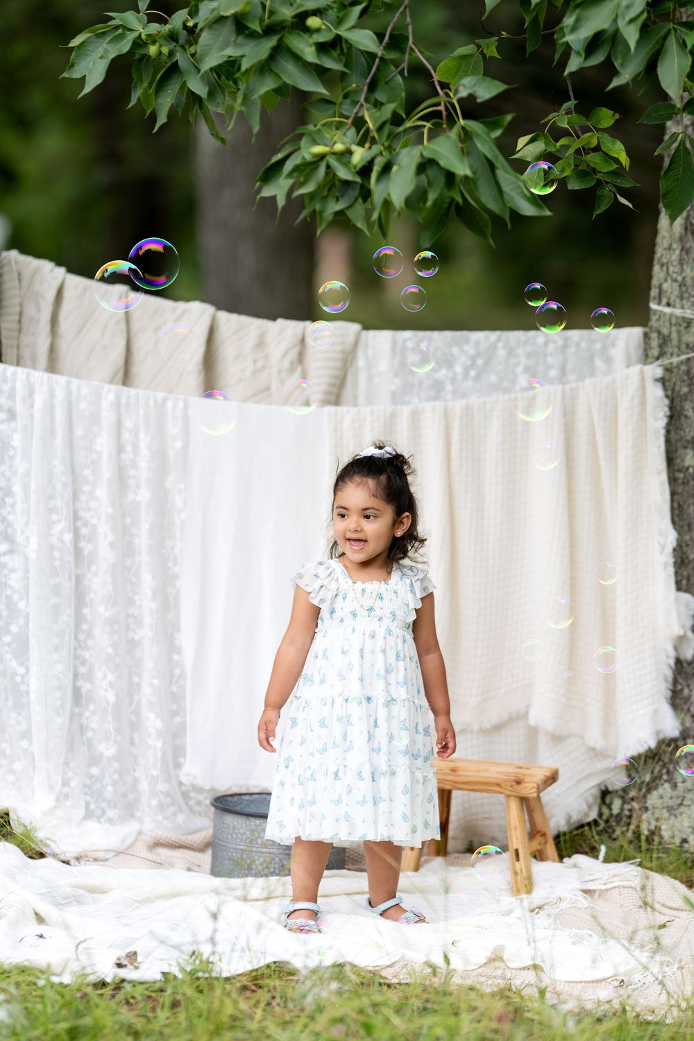 Young girl in white dress stands outdoors near clothesline with bubbles floating around her in a lush green garden.