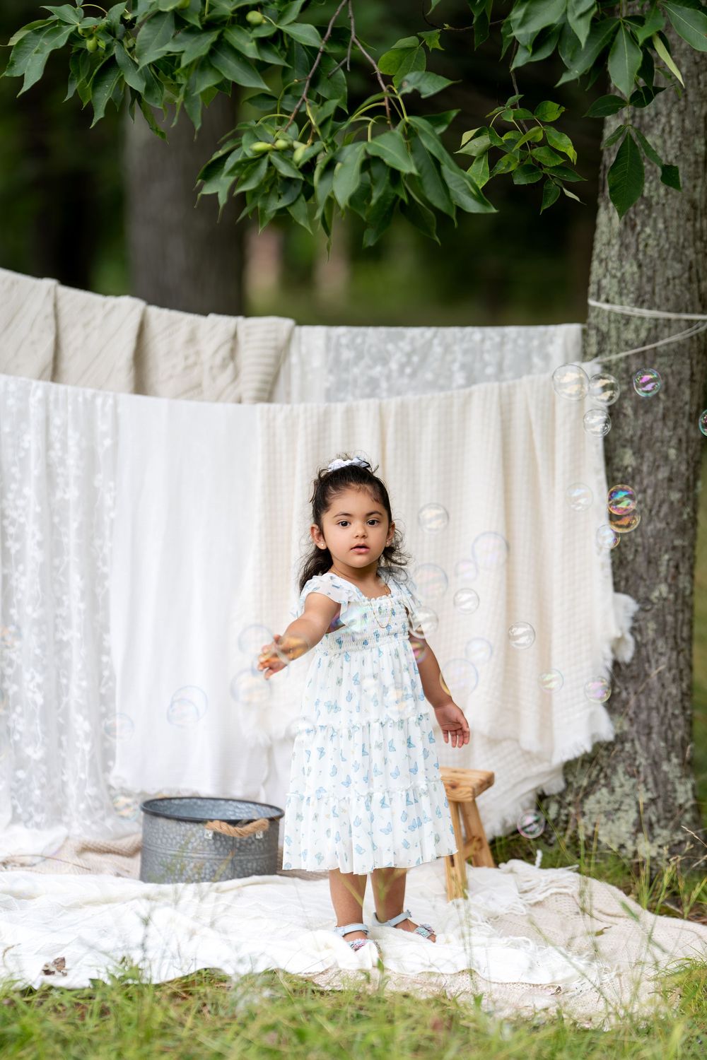 Young girl in floral dress playing with bubbles outdoors near a clothesline with white linens hanging.