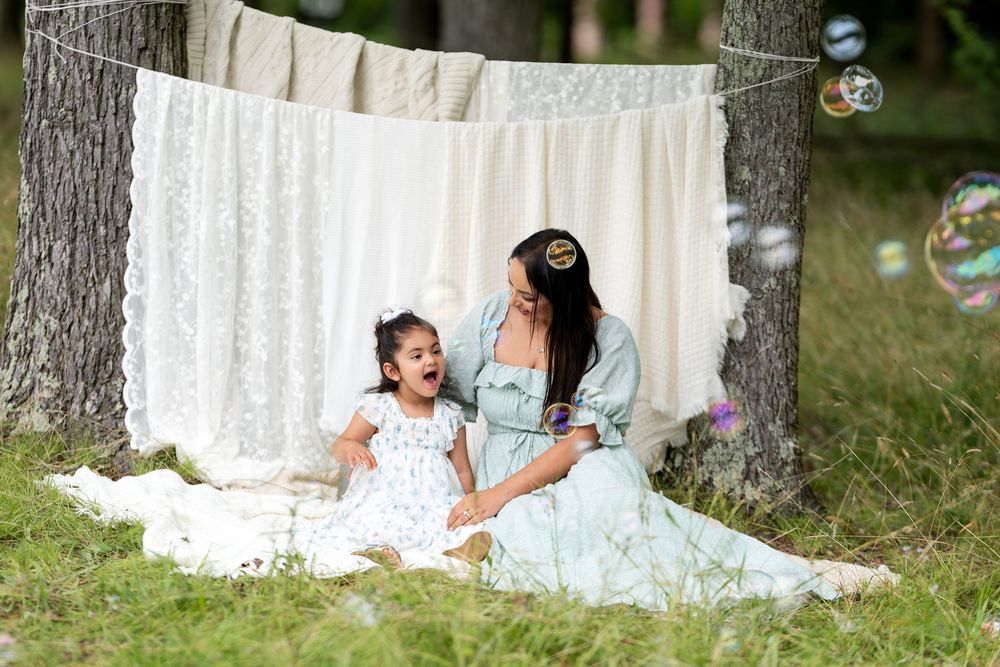 Mother and daughter in matching dresses laugh together on a blanket outdoors with bubbles and fabric backdrop.